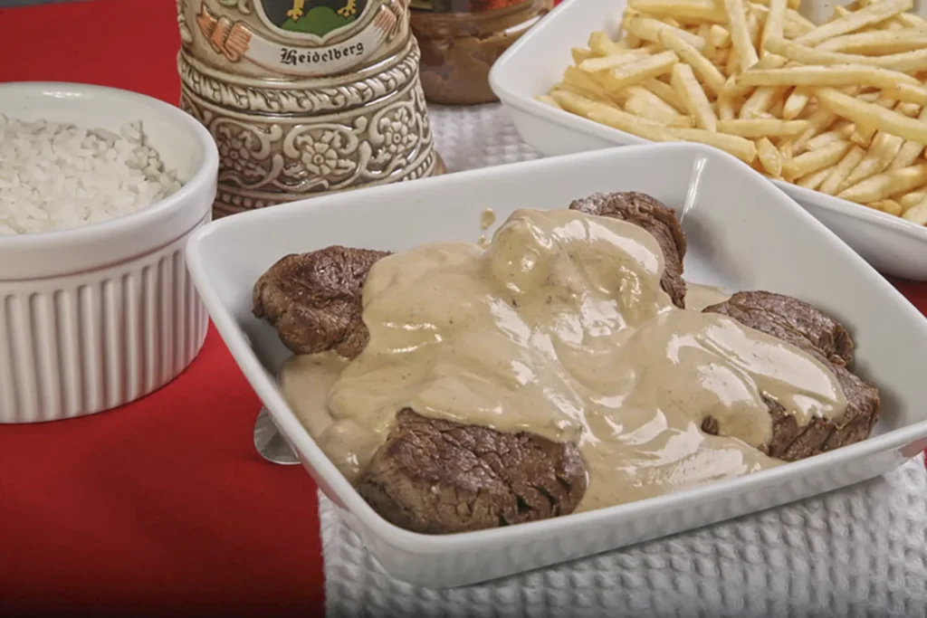 Medalhões de filé-mignon cobertos por um farto e cremoso molho de mostarda, servidos em uma travessa branca. O prato é acompanhado por porções de arroz branco e batatas fritas, com um tradicional caneco de chope alemão ao fundo.