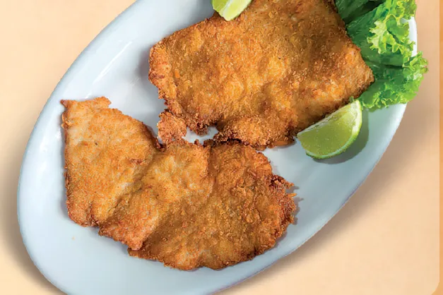 Prato típico alemão de Schnitzel, composto por dois grandes filés de carne finamente batidos e empanados, fritos até atingirem uma cor dourada e crocante, servidos em uma travessa branca com fatias de limão fresco e alface.